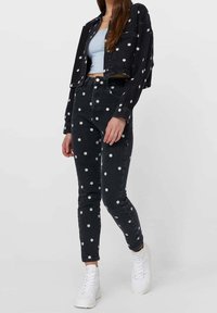 Czarna, dżinsowa kurtka z krótkim rękawem i spodnie z wysokim stanem w białe wzory stokrotek, zestawione z białymi adidasami. Gładka tekstura i dopasowany fason.