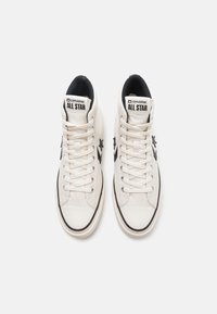 Para białych sneakersów typu high-top Converse All Star z czarnym logo gwiazdy i białymi sznurowadłami na jednolitym jasnym tle.