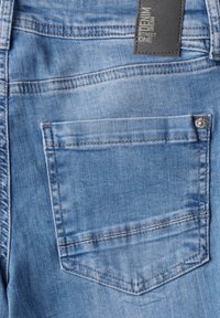 Primer plano de unos jeans de mezclilla azul claro, con un bolsillo trasero que presenta costuras en la parte superior y una etiqueta de tela unida a la cinturilla.