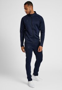 Marine trainingspak gemaakt van gladde, rekbare stof. Bevat een zipjack met staande kraag en bijpassende, taps toelopende broek, beide met logoaccenten.