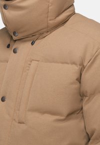 Hnědá péřová bunda s vysokým límcem, prošívanou texturou a cvočkovými detaily. Obsahuje zipovou náprsní kapsu a nastavitelnou šňůrku.