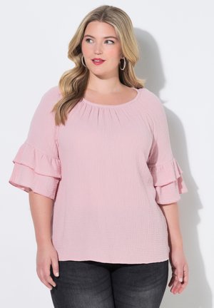 Kvinde med langt blondt hår iført en lyserød tekstureret bluse med lagdelte flæseærmer og mørke jeans, der kigger let til siden.