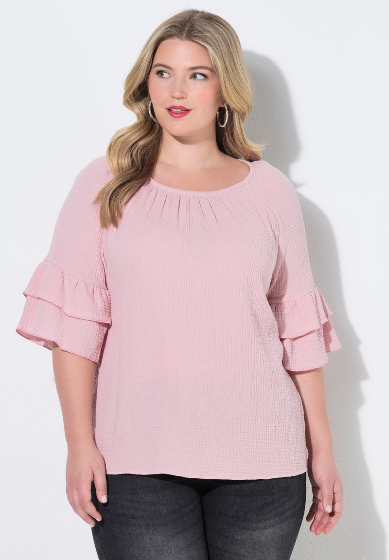 Mujer de cabello largo y rubio que lleva una blusa rosa con textura y mangas con volantes en capas, y vaqueros oscuros, mirando ligeramente hacia un lado.