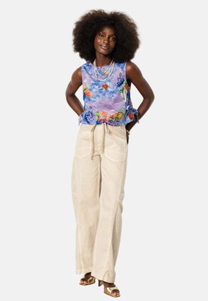 Femme aux cheveux longs et bouclés portant un haut violet sans manches à motif floral, un pantalon beige à jambes larges, des colliers superposés et des sandales dorées à talons.
