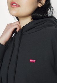 Femme portant un sweat à capuche noir Levi's, tenant le col près de son cou, montrant un petit logo rouge Levi's sur la poitrine.