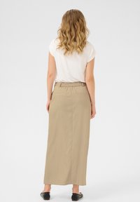 Jupe maxi beige avec une taille élastique, deux poches arrière et une texture lisse. Portée avec un haut blanc à manches courtes et des ballerines noires.