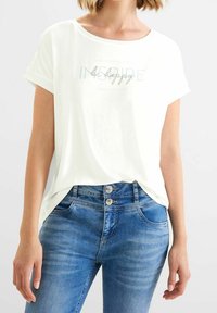 Vit t-shirt med kort ärm och rund halsringning, prydd med ljusblå text och dekorationer, tillsammans med mid-blå denimjeans.