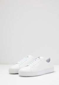 Bílé kožené tenisky s kulatými špičkami, plochými tkaničkami a texturovanými gumovými podešvemi, které mají minimalistický design a žádná viditelná loga.