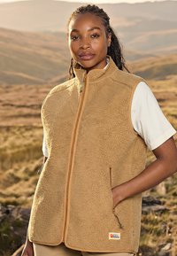 Jeune femme aux cheveux tressés portant un gilet en polaire beige par-dessus un t-shirt blanc, debout en plein air dans un paysage vallonné au coucher du soleil.