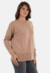 Una donna con capelli castani di lunghezza spalla, che indossa un maglione beige a maniche lunghe e jeans scuri, in piedi di fronte a uno sfondo bianco.