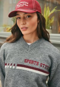 Jeune femme portant une casquette rouge et un pull gris avec le texte "SPORTS STUDIO", se tenant à l'intérieur près de plantes et de fenêtres.