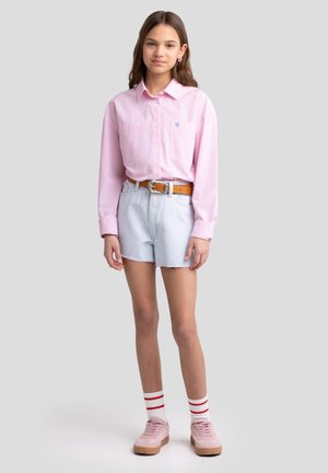 Hellrosa gestreifte Bluse mit Knöpfen und zwei Brusttaschen, kombiniert mit hellblauen Denimshorts. Brauner Gürtel, rosa Turnschuhe und gestreifte weiße Socken.