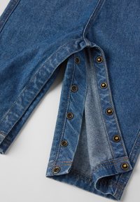 Denim broek met een donkerblauwe wassing, voorzien van koperen drukknopen langs de zoom, versterkte stiksels en een iets uitgescheurde binnennaad.