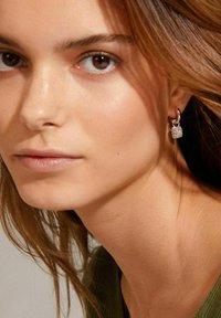 Boucles d'oreilles en argent avec un charm rectangulaire texturé et scintillant. Gros plan d'un visage avec un maquillage léger et de longs cheveux bruns.