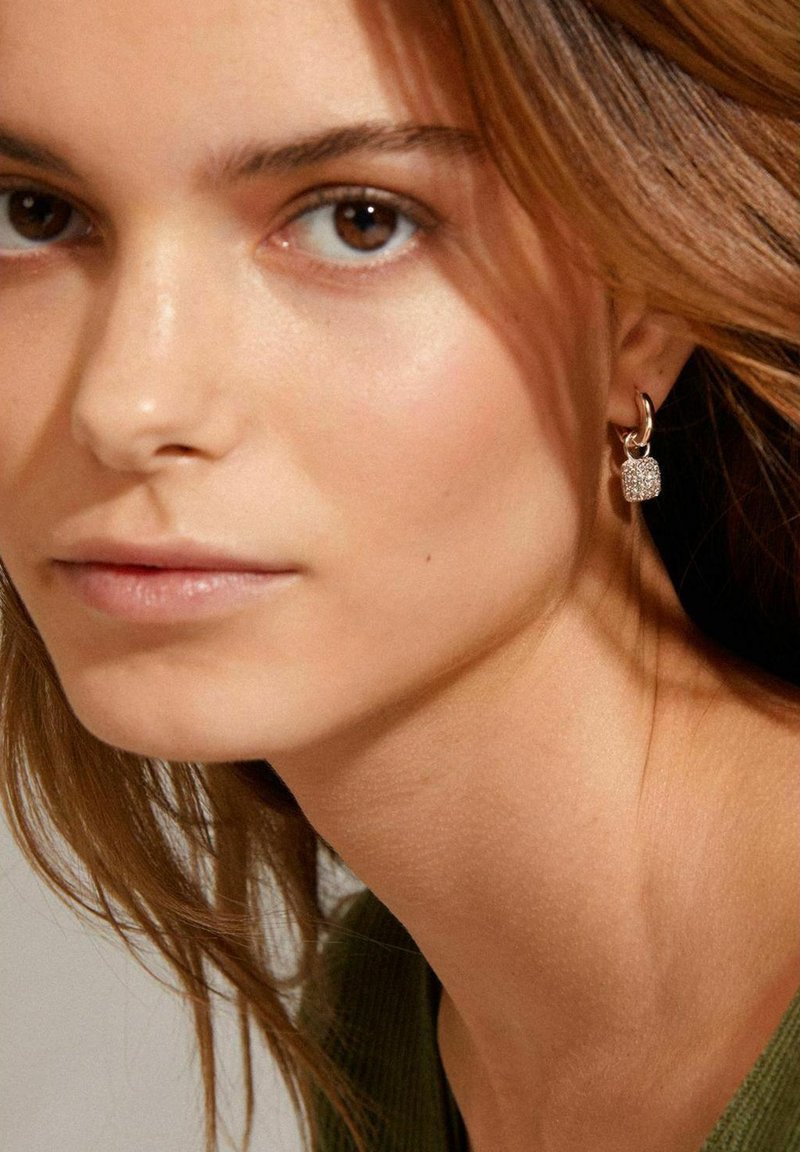Boucles d'oreilles en argent avec un charm rectangulaire texturé et scintillant. Gros plan d'un visage avec un maquillage léger et de longs cheveux bruns.