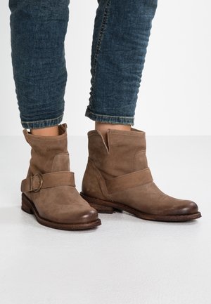 Bottines en cuir marron avec boucle latérale portées avec un jean bleu retroussé sur un sol blanc.