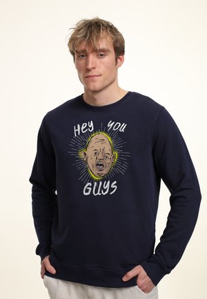 Mand med totet blondt hår iført navy sweatshirt med en karikeret skaldet ansigt og teksten "Hey you guys" i hvid.
