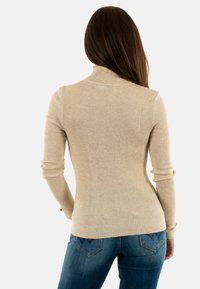 Beige gerippter Rollkragenpullover mit langen Ärmeln, figurbetontem Design und subtiler Textur. Kombiniert mit blauen Jeans mit dekorativen Gesäßtaschen.