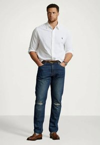 Bílá košile na knoflíky, modré džíny s opranými detaily, hnědý opasek, hnědé boty a stříbrné hodinky. Muž stojí na neutrálním pozadí.