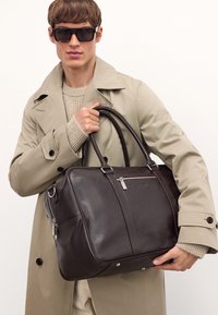 Mørkebrunt læderhåndtaske med dobbelte håndtag, sølvhardware og en frontlomme med lynlås. Glat tekstur, rektangulær form.
