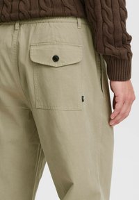 Beige cotton trousers with side pockets, back flap pocket with black button, and a relaxed fit; textured fabric, visible stitching details.