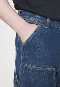 Dunkelblaue Denim-Cargohosen mit einer Seitentasche, sichtbaren Nähdetails und einer strukturierten Oberfläche, mit einer Nahaufnahme des Bundes.