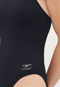 Nahaufnahme einer Person, die einen schwarzen Speedo Endurance+-Badeanzug trägt, mit Fokus auf das Logo und den seitlichen Torsobereich.