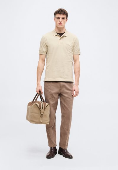 Jeune homme portant un polo beige et un pantalon marron, tenant un sac de voyage beige et marron, debout devant un fond gris clair.