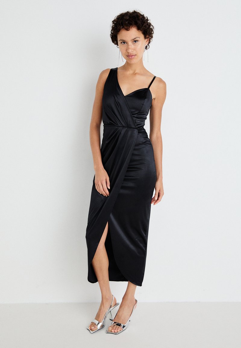 Abito in satin nero con design a una spalla, parte anteriore incrociata e spacco fino alla coscia. Modella che indossa sandali argentati con tacco.
