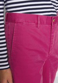 Pantaloni di cotone rosa con una texture liscia, dotati di chiusura con bottone e tasche laterali, abbinati a una camicia a maniche lunghe a righe blu e bianche.