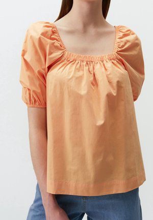 Femme portant un chemisier ample couleur pêche avec des manches bouffantes, un décolleté carré froncé et un jean bleu.