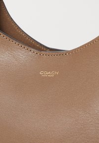 Geantă din piele maro cu textură netedă, având un logo gravat cu aur "Coach New York" și detalii de cusătură pe margini.
