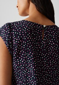 Blusa navy con motivo a pois in rosa e bianco. Presenta una chiusura a chiave sul retro con un bottone e maniche corte, leggermente a sbuffo.