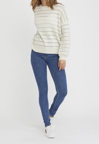 Suéter crema claro con rayas plateadas horizontales; escote redondo y mangas abullonadas. Combinado con jeans ajustados azul oscuro y zapatillas blancas.