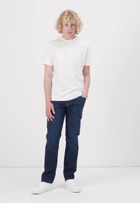 Hvit bomulls t-skjorte med "GUESS" logo, kombinert med mørkeblå denim jeans og hvite sneakers. Rett passform med myk tekstur.
