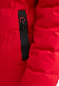 Primer plano de la manga de una chaqueta acolchada roja con detalle de correa negra y un lazo de cordón.