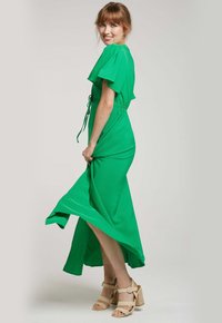 Robe longue verte avec de courtes manches volantées, taille cintrée et jupe fluide. Portée avec des sandales légères à lanières. Texture de tissu lisse.