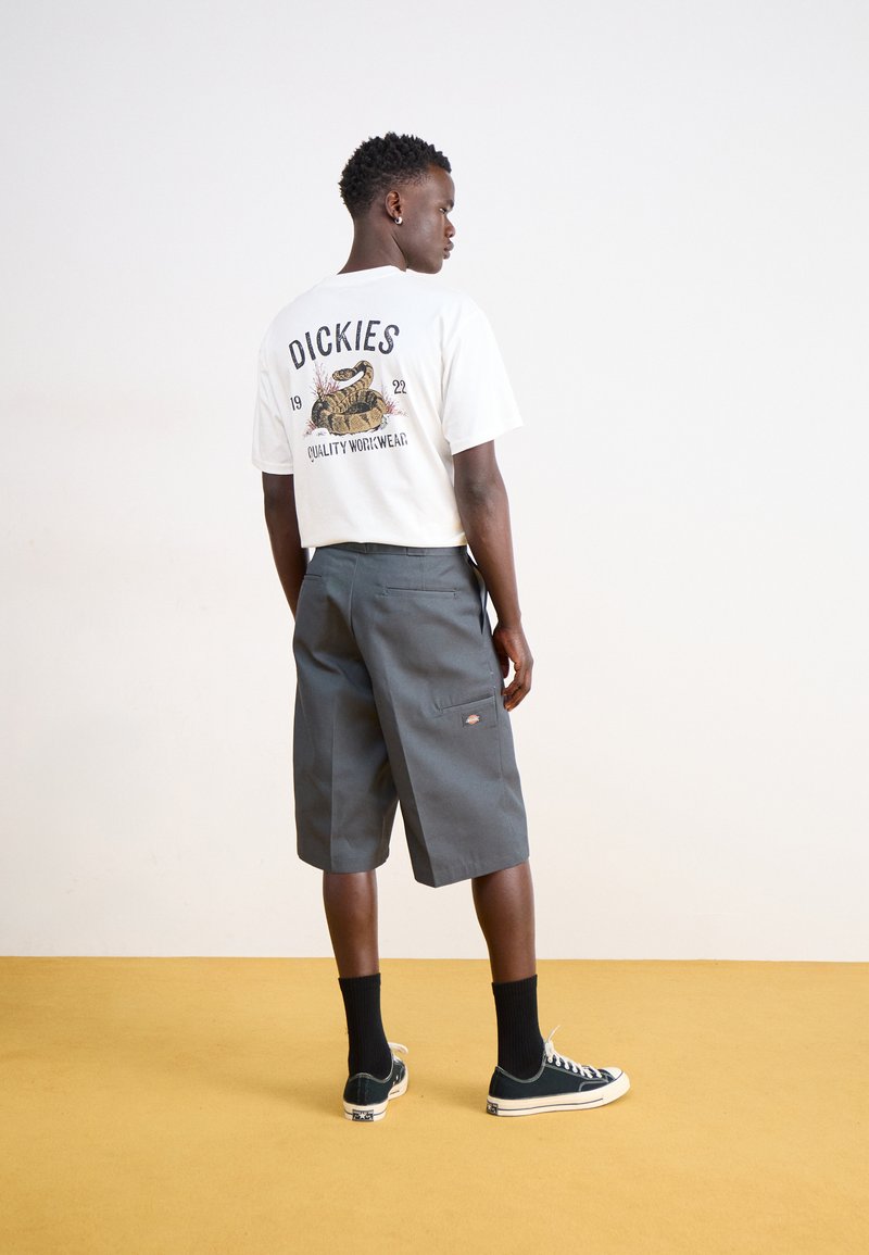 T-shirt bianca con una grafica di un serpente e il testo "DICKIES" sul retro. Pantaloni corti grigi con tasche e calzini neri con sneakers verdi su un pavimento giallo.
