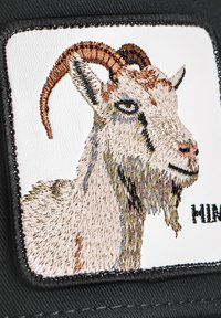 Embroidered patch of a goat's head on a white background, bordered by black fabric; details include brown horns and textured fur.