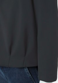 Zwarte blouse met een zachte, gladde textuur en lange mouwen; heeft een subtiele plooi bij de zoom; gecombineerd met blauwe jeans.