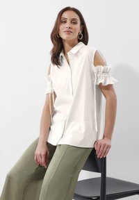 Witte korte mouwen blouse met uitsnijdingen op de schouders en ruffles. Gemaakt van een gladde stof, met een knoopsluiting aan de voorkant. Gecombineerd met olijfgroene broek.