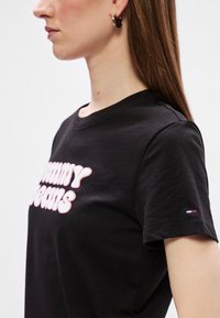 Μαύρο βαμβακερό T-shirt με έντονη εκτύπωση "Tommy Jeans" σε ροζ. Κοντά μανίκια με μικρό τριχρωμο λογότυπο στον αριστερό ώμο.