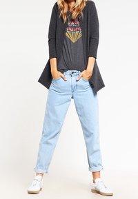 Grå långärmad cardigan över en graphic t-shirt med flerfärgad text, matchad med ljusblå upprullade jeans och vita sneakers.