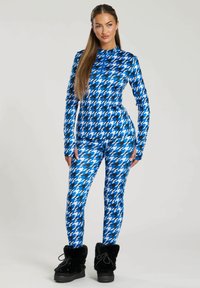 Vrouw in een lange mouw top met houndstooth patroon in blauw, zwart en wit, en legging, met zwarte winterlaarzen met bontvoering.