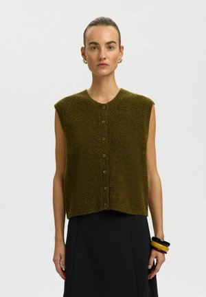 Femme aux cheveux plaqués en arrière portant un gilet en tricot boutonné sans manches vert olive, une jupe noire, des boucles d'oreilles créoles dorées et des bracelets noirs et jaunes.