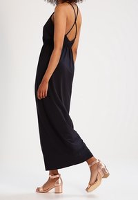 Schwarzes, ärmelloses Maxikleid mit einer taillierten Passform und dünnen, überkreuzten Trägern am Rücken. Kombiniert mit metallicfarbenen, Blockabsatzsandalen.
