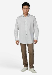 Ein Mann steht mit dem Gesicht nach vorne, trägt ein hellgemustertes Hemd mit Knöpfen, braune Hosen und weiße Turnschuhe vor einem schlichten weißen Hintergrund.