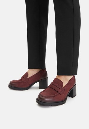 Vīna sarkana zamša loaferi ar bloka papēdi, ar diviem priekšējiem siksniņām un gludu ādas apmali, kombinēti ar pielāgotām melnām biksēm.