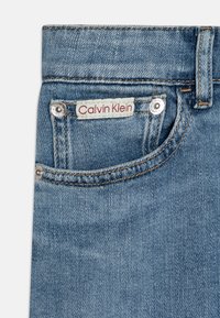 Jasnoniebieskie dżinsy z górną kieszenią z czerwoną metką "Calvin Klein", srebrnymi nitami oraz kontrastującym pomarańczowym przeszyciem.