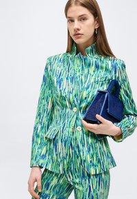 Jeune femme portant un costume à motifs verts et bleus tenant un petit sac à main bleu foncé, faisant face à l'avant avec une expression neutre.